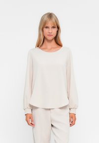 Ανοιχτό καφέ μπλούζα μακρύ μανίκι με στρογγυλή λαιμόκοψη και ελαστικούς καρπούς, φτιαγμένη από λείο ύφασμα, σε συνδυασμό με αντίστοιχες ανοιχτόχρωμες παντελόνες.