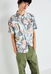 Kurzarm-Hemd mit Knopfleiste in einem bunten abstrakten Muster aus Pastellrosa, Blau und Grau, kombiniert mit olivgrünen Hosen.