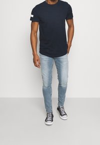 Camiseta de algodón azul marino con mangas cortas y una franja blanca en el hombro, combinada con unos jeans fit de color azul claro y zapatillas negras.