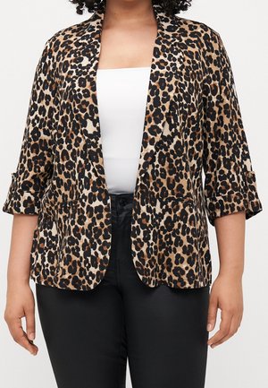 Femme portant un blazer à imprimé léopard avec les manches retroussées sur un top blanc et un pantalon noir, sur un fond uni.
