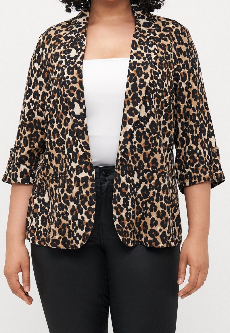 Femme portant un blazer à imprimé léopard avec les manches retroussées sur un top blanc et un pantalon noir, sur un fond uni.