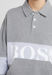 Camisa polo gris con cuello, tres botones blancos y un panel blanco con la palabra "BOSS" mostrada de manera destacada. Textura suave.