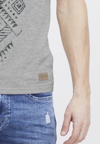 Graues T-Shirt mit schwarzem geometrischem Druck auf der Vorderseite. Verfügt über ein kleines braunes Etikett am Saum und wird mit abgewetzten blauen Jeans kombiniert.
