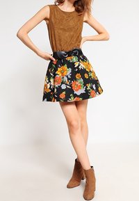 Braun ärmelloses Oberteil kombiniert mit einem schwarzen Blumenrock, der orangefarbene und gelbe Blumen zeigt; unterstrichen mit einem schwarzen Gürtel. Braune Knöchelstiefel.