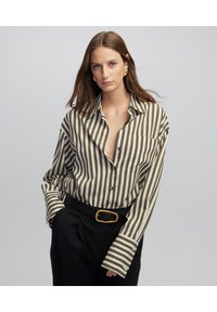 Camicia a righe color verde oliva e crema, con una vestibilità morbida, scollo a V profondo e maniche lunghe con ampi polsini, abbinata a pantaloni neri.