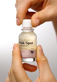 Bouteille en verre transparent avec un bouchon blanc, remplie d'un liquide rose clair. L'étiquette contient le nom du produit et les ingrédients. Une main saisit la bouteille.