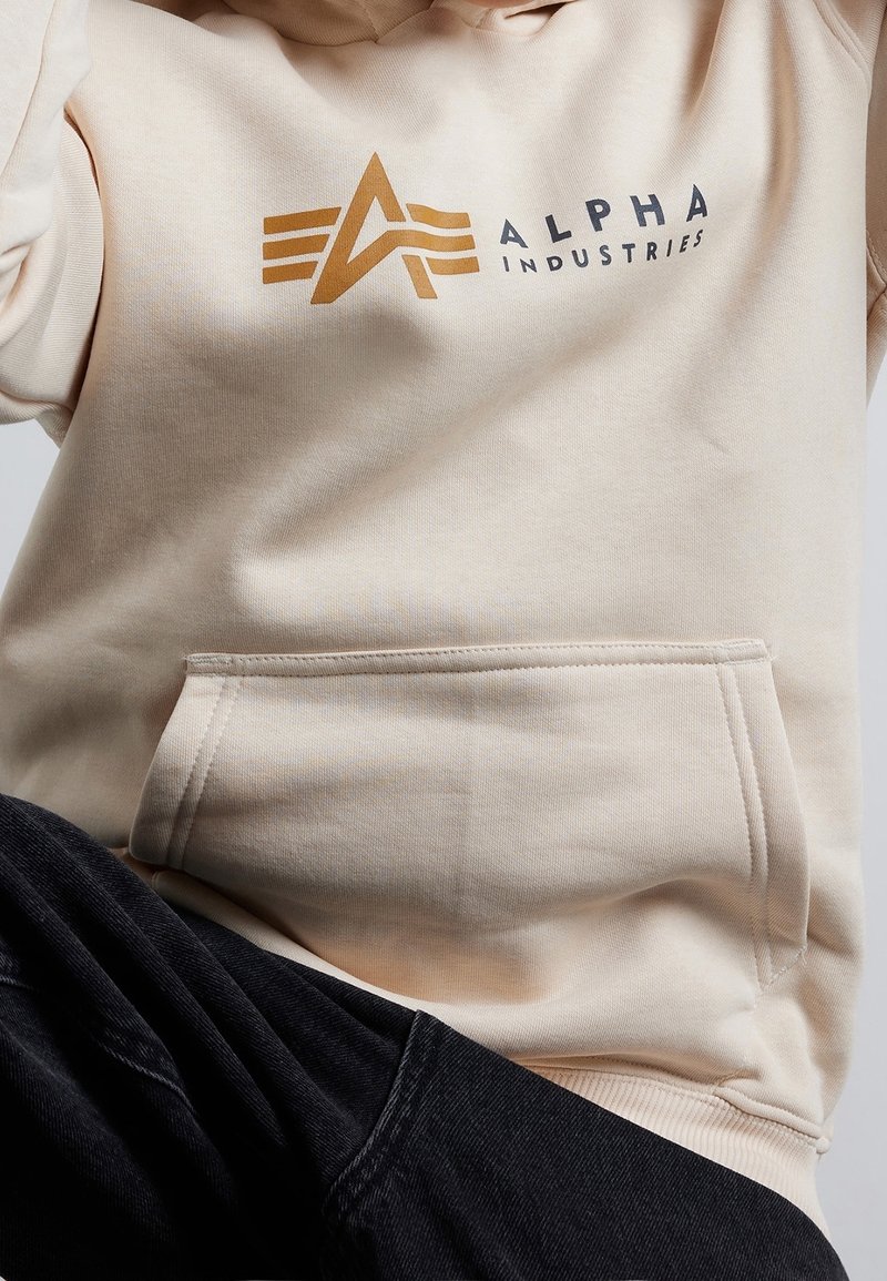 Hoodie beige con logo e scritte dorate, tessuto morbido, tasca frontale e polsini a coste. Abbinato a pantaloni scuri.
