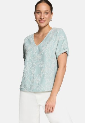 Femme portant un haut à manches courtes bleu clair à col en V avec une broderie florale blanche et un pantalon blanc, souriante, avec la main détendue sur le côté.