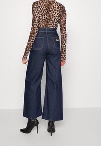 Jeans de mezclilla oscuros de tiro alto y pierna ancha, con dos bolsillos traseros y detalles de costura, combinados con una blusa transparente de leopardo de manga larga.
