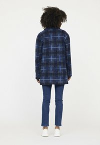 Blauwe geruite jas van dik materiaal, met een boxy ontwerp en een kraag. Gecombineerd met aansluitende spijkerjeans en witte sneakers.