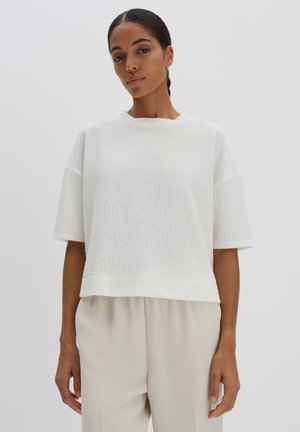 Textured white knit top with short sleeves, round neck, and a loose fit, paired with light beige trousers. Smooth fabric and relaxed shape.