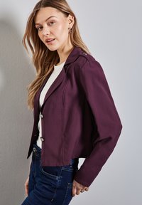 Giacca cropped viola scuro realizzata in tessuto liscio, con colletto a punta e bottoni argentati, abbinata a una maglietta bianca e jeans blu.