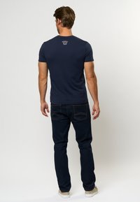 Mørkeblå tettsittende t-skjorte, kort ermer, ribbestrikket hals, med hvit logo på ryggen; parret med mørk denim jeans og lyse sneakers.