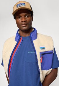 Patagonia vest in crèmekleurige fleece met blauwe accenten, met rits en zak. Blauwe t-shirt eronder met groen logo, en tankleurige pet met logo.