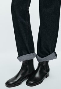 Bottes noires en cuir portées avec un jean foncé retroussé, sur un fond clair uni.