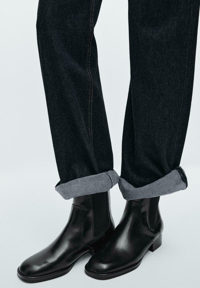 Bottes noires en cuir portées avec un jean foncé retroussé, sur un fond clair uni.