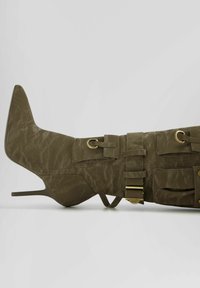 Olivgrön stilettkänga med strukturerad yta, sidbucklor och spetsig tå. Har en hög klack och dekorativa remmar.