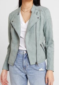 Veste en cuir synthétique vert menthe avec une coupe courte, des cols style motard, des poches zippées et des détails côtelés sur les épaules.