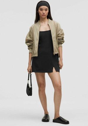 Giacca bomber beige, mini abito nero con spacco laterale, scarpe slip-on nere, con una piccola borsa nera, su sfondo neutro.