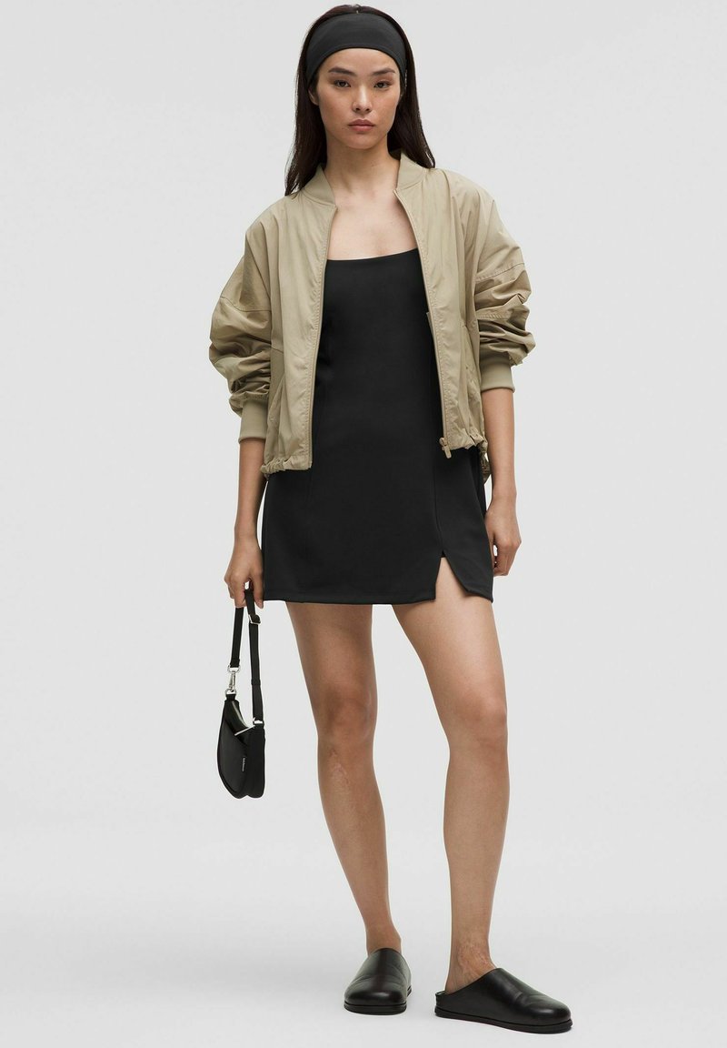Chaqueta bomber beige, vestido mini negro con abertura lateral, zapatos negros sin cordones, sosteniendo una pequeña bolsa negra, sobre un fondo neutro.