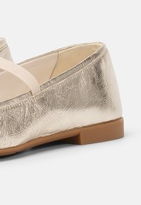 Metallic gold ballet flat with a smooth, textured finish, flexible rubber sole, and a soft, contrasting elastic strap across the front.
