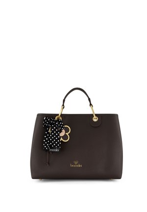 Borsa a mano in pelle marrone scuro con dettagli in oro, un fiocco a pois bianco e nero e un ciondolo dorato a forma di fiore appeso a una maniglia.