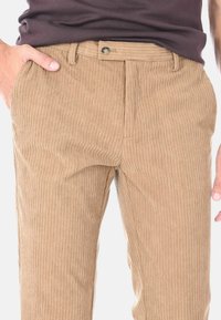 Uomo che indossa pantaloni in corduroy beige con righe verticali, chiusura a bottoni e mano nella tasca sinistra, abbinati a una camicia marrone scuro.