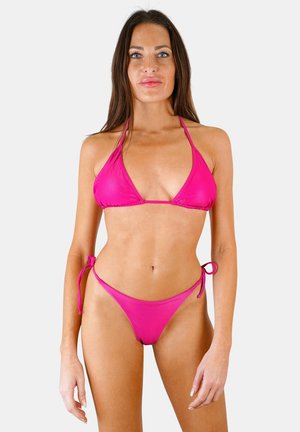 Femme debout face à l'avant portant un bikini triangle rose vif avec un bas noué sur les côtés sur un fond clair uni.