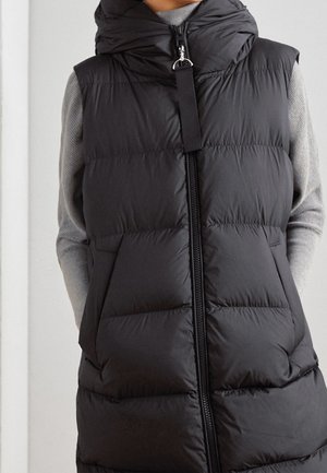 Doudoune sans manches noire matelassée avec fermeture éclair frontale et capuche, portée sur un pull gris clair à manches longues.