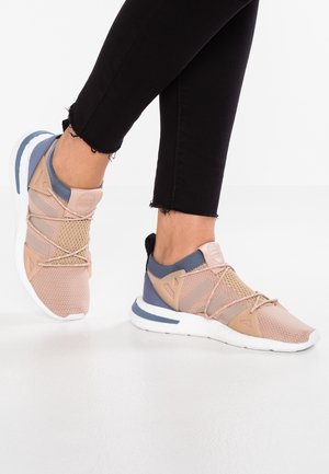 Person iført pink og blå sporty sneakers med hvide såler, kombineret med sorte ankel-lange bukser på et hvidt gulv.