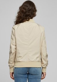 Bézs bombázó dzseki sima felülettel, bordázott mandzsettákkal és szegéllyel, valamint texturált oldalsó panelekkel. Egyszerű kialakítású, látható záródások nélkül.