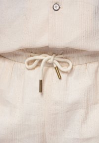 Gros plan sur un pantalon en tissu beige avec un cordon de serrage noué comportant des embouts métalliques et une partie d'une chemise assortie avec un bouton visible.