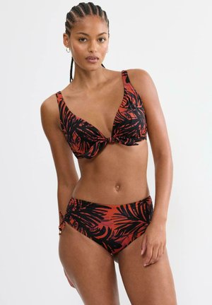 Frau mit geflochtenem Haar trägt einen Bikini mit tropischem Blattmuster in Rot und Schwarz, mit Knotendetail am Oberteil und gerafften Seiten am Unterteil.