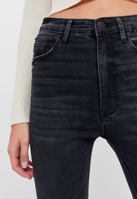 Jeans de mezclilla negra con un diseño ajustado, que cuentan con bolsillos frontales, pasadores para cinturón y una textura sutil. La tela tiene un ligero brillo.