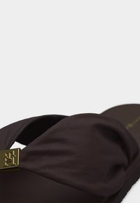 Παντόφλα σε σκούρο καφέ με πλατιά υφασμάτινη λωρίδα που συγκεντρώνεται σε ένα χρυσό τετραγωνικό έμβλημα πάνω από το πόδι, με ομαλή σόλα που φαίνεται στο πλάι.