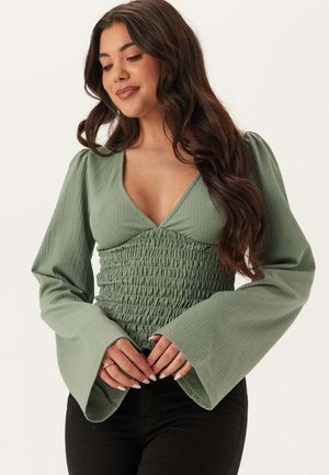 Kvinna med långt mörkt hår som bär en grön texturerad långärmad topp med rynkad midja och utsvängda ärmslut, poserar med händerna knäppta.