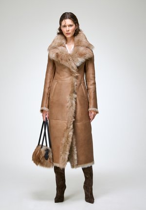 Femme portant un long manteau en cuir marron avec garniture en fourrure, tenant un sac à main en fourrure assorti, et portant des bottes marron foncé.