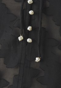 Blusa in tessuto nero trasparente con texture floreale, caratterizzata da diverse perle irregolari e hardware dorato lungo la chiusura centrale.