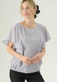 Una donna con i capelli biondi raccolti da un elastico nero indossa orecchini a cerchio dorati e collane a strati, una blusa leggera di colore viola chiaro con maniche a sbuffo e pantaloni neri.