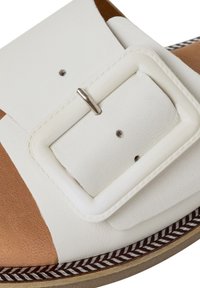 Gros plan d'une sandale en cuir blanc avec une grande boucle carrée et une zone des orteils marron sur le bord d'une semelle à motif.