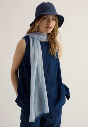 Nő sötétkék ujjatlan felsőben és nadrágban, világoskék sállal és sötétkék kalapban, egyszínű világos háttér előtt áll.