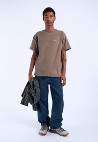 T-shirt in cotone marrone con un dettaglio grafico, jeans a gamba larga blu e camicia di flanella a fantasia in mano. Sneakers grigie e beige con accenti.