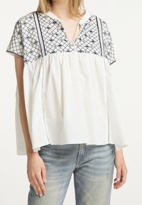 Blouse blanche à manches courtes avec un empiècement à motifs bleu marine, un décolleté en V et un corps ample froncé avec des coutures texturées.