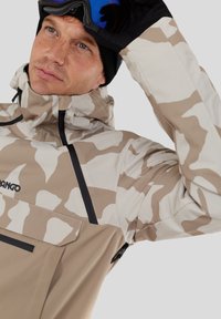 Beige und weiße Camouflage-Skijacke mit hohem Kragen, Reißverschlusstasche und schwarzen Akzenten. Glatte Textur und wasserabweisendes Material.
