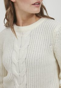 Hvit strikket genser med et kabelstrikkedesign i midten, ribbet rund hals og en åpen vevstruktur gjennom hele genseren.