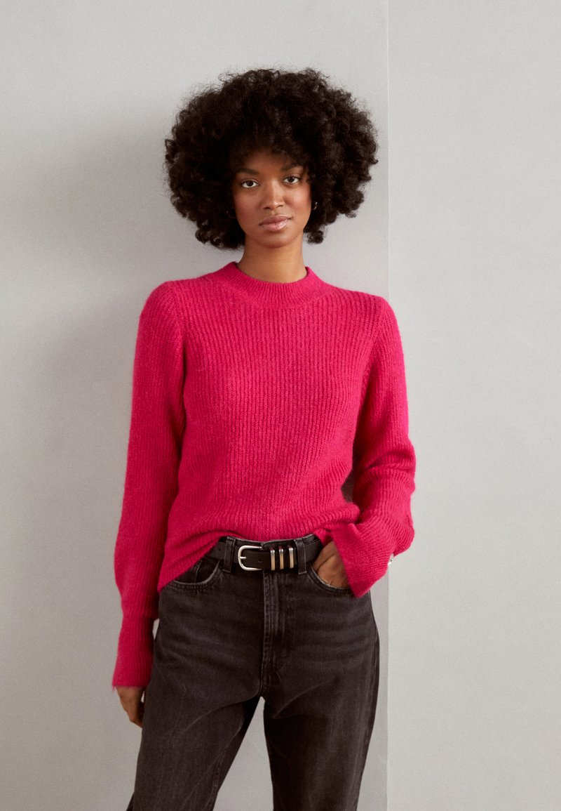 Roze gebreide trui met geribbelde textuur, ronde halslijn en lange mouwen, gecombineerd met hooggewaisted donkere denim jeans en een zwarte riem.