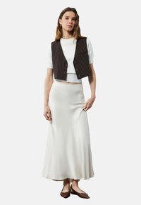 Chaleco marrón de rayas sobre una blusa blanca de manga corta y una falda larga de satén blanca, combinada con zapatos planos marrones.