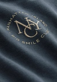 T-shirt i bomull, marinblå, med broderad logotyp "NYC" i krämfärg, med texten "Manhattan Island Big Smile Club" runt designen.