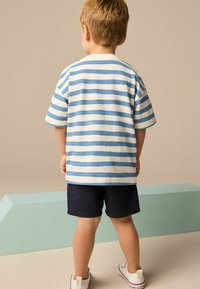 Gestreiftes Baumwoll-T-Shirt in Blau und Weiß, kurze Ärmel, kombiniert mit marineblauen Shorts. Das Kind trägt weiße Sneaker mit einem Rückenzugdetail.