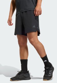 Pantalones cortos deportivos negros con tela ligera, que presentan un logotipo de Adidas y un dobladillo dividido. Combinados con calcetines negros y zapatillas de malla negras.